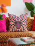 Square psychedelic folk art throw pillow cover printed to look like textured embroidery on a cream background. A large black spider with coral red and magenta markings on its legs and abdomen sits at the center, surrounded by rippling waves in blue violet, coral red, magenta, and golden yellow. Smooth flat 2D print, not raised stitching. Retro gothic Halloween maximalist decor for a sofa, armchair, or goth bedroom, made for spooky season.