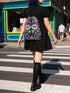 Drawstring backpack measuring 15 by 17 inches, made of 100% polyester with narrow shoulder straps and a drawstring closure. A symmetrical pastel goth print of bats, stylized eyes, flowers, and stars in a soft pastel palette sits on a black base, framed by purple and green checkerboard bands. Creepy cute, whimsigoth, kawaii goth style bag made for the gym, concerts, festivals, travel, school, and everyday carry.