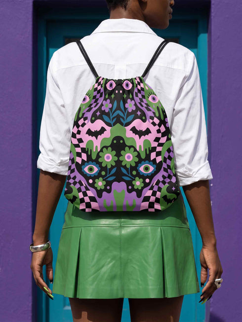 Drawstring backpack measuring 15 by 17 inches, made of 100% polyester with narrow shoulder straps and a drawstring closure. A symmetrical pastel goth print of bats, stylized eyes, flowers, and stars in a soft pastel palette sits on a black base, framed by purple and green checkerboard bands. Creepy cute, whimsigoth, kawaii goth style bag made for the gym, concerts, festivals, travel, school, and everyday carry.