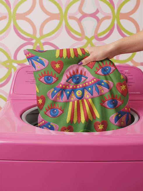 Square weirdcore throw pillow cover printed to look like textured crewel embroidery on a green background. A pink and blue flying saucer with one large blue eye at its center hovers among floating blue eyeballs and red hearts. Smooth flat 2D print, not raised stitching. Maximalist, trippy sci-fi dopamine decor with quirky alien charm for a sofa, dorm bed, gaming chair, or reading nook, made for space lovers and sci-fi fans.