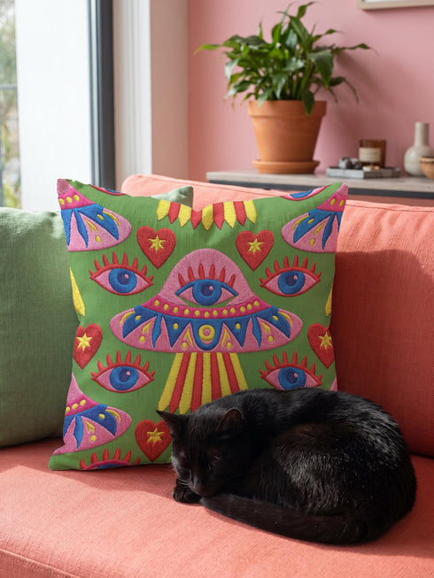 Square weirdcore throw pillow cover printed to look like textured crewel embroidery on a green background. A pink and blue flying saucer with one large blue eye at its center hovers among floating blue eyeballs and red hearts. Smooth flat 2D print, not raised stitching. Maximalist, trippy sci-fi dopamine decor with quirky alien charm for a sofa, dorm bed, gaming chair, or reading nook, made for space lovers and sci-fi fans.