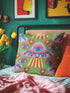 Square weirdcore throw pillow cover printed to look like textured crewel embroidery on a green background. A pink and blue flying saucer with one large blue eye at its center hovers among floating blue eyeballs and red hearts. Smooth flat 2D print, not raised stitching. Maximalist, trippy sci-fi dopamine decor with quirky alien charm for a sofa, dorm bed, gaming chair, or reading nook, made for space lovers and sci-fi fans.
