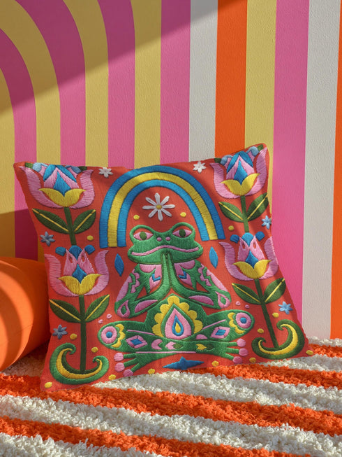 Square folk art throw pillow cover printed to look like textured crewel embroidery on a coral orange background. A green frog sits cross-legged in a meditation pose beneath a blue and yellow arch, surrounded by symmetrical pink flowers. Smooth flat 2D print, not raised stitching. Colorful, whimsical, maximalist dopamine decor for a yoga room, meditation corner, sofa, or reading nook, made for yoga lovers and frog fans.