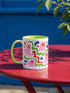 Glossy white ceramic 11 oz coffee mug with a lime green rim, inside, and handle and a full wraparound pastel goth print. Pink ghosts with paired red oval eyes and rounded scalloped lower edges float between wavy green and red ribbons. Blue starbursts, curved blue shapes, red teardrops, small colorful dots, and green diamond sparkles fill the white background. Creepy cute Halloween coffee mug with a playful, retro maximalist feel.