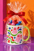 Glossy white ceramic 11 oz coffee mug with a lime green rim, inside, and handle and a full wraparound pastel goth print. Pink ghosts with paired red oval eyes and rounded scalloped lower edges float between wavy green and red ribbons. Blue starbursts, curved blue shapes, red teardrops, small colorful dots, and green diamond sparkles fill the white background. Creepy cute Halloween coffee mug with a playful, retro maximalist feel.