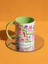 Glossy white ceramic 11 oz coffee mug with a lime green rim, inside, and handle and a full wraparound pastel goth print. Pink ghosts with paired red oval eyes and rounded scalloped lower edges float between wavy green and red ribbons. Blue starbursts, curved blue shapes, red teardrops, small colorful dots, and green diamond sparkles fill the white background. Creepy cute Halloween coffee mug with a playful, retro maximalist feel.