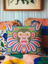 Square folk art throw pillow cover printed to look like thick crewel yarn embroidery. A bold, whimsical monkey face in olive green and pink stares out with intense yellow eyes, framed by a retro scalloped floral collar. Smooth flat 2D print, not raised stitching. Clowncore, junglecore, and weirdcore maximalist decor with eclectic, quirky charm for a sofa, accent chair, or bedroom, made for animal lovers and alt renters.