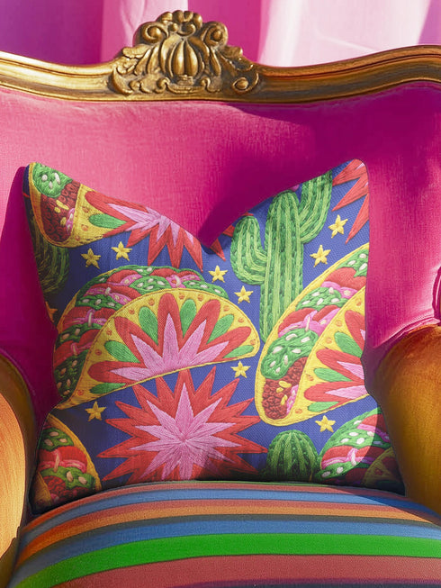 Square maximalist throw pillow cover printed to look like bold crewel embroidery on a solid royal blue background. Tacos with green, red, and brown fillings scatter across the design with bright green saguaro cacti, large pink and red starburst flowers, and small yellow stars. Smooth flat 2D print, not raised stitching. Colorful, quirky dopamine decor for a sofa, kitchen bench, or breakfast nook, made for taco lovers and foodies.