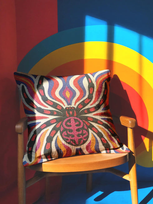 Square psychedelic folk art throw pillow cover printed to look like textured embroidery on a cream background. A large black spider with coral red and magenta markings on its legs and abdomen sits at the center, surrounded by rippling waves in blue violet, coral red, magenta, and golden yellow. Smooth flat 2D print, not raised stitching. Retro gothic Halloween maximalist decor for a sofa, armchair, or goth bedroom, made for spooky season.