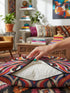 Square psychedelic folk art throw pillow cover printed to look like textured embroidery on a cream background. A large black spider with coral red and magenta markings on its legs and abdomen sits at the center, surrounded by rippling waves in blue violet, coral red, magenta, and golden yellow. Smooth flat 2D print, not raised stitching. Retro gothic Halloween maximalist decor for a sofa, armchair, or goth bedroom, made for spooky season.
