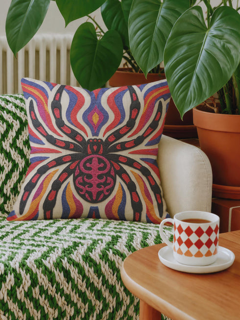 Square psychedelic folk art throw pillow cover printed to look like textured embroidery on a cream background. A large black spider with coral red and magenta markings on its legs and abdomen sits at the center, surrounded by rippling waves in blue violet, coral red, magenta, and golden yellow. Smooth flat 2D print, not raised stitching. Retro gothic Halloween maximalist decor for a sofa, armchair, or goth bedroom, made for spooky season.
