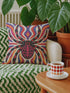 Square psychedelic folk art throw pillow cover printed to look like textured embroidery on a cream background. A large black spider with coral red and magenta markings on its legs and abdomen sits at the center, surrounded by rippling waves in blue violet, coral red, magenta, and golden yellow. Smooth flat 2D print, not raised stitching. Retro gothic Halloween maximalist decor for a sofa, armchair, or goth bedroom, made for spooky season.
