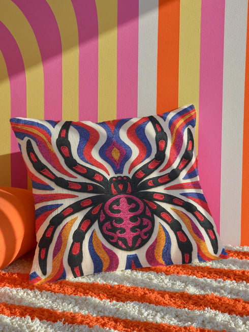 Square psychedelic folk art throw pillow cover printed to look like textured embroidery on a cream background. A large black spider with coral red and magenta markings on its legs and abdomen sits at the center, surrounded by rippling waves in blue violet, coral red, magenta, and golden yellow. Smooth flat 2D print, not raised stitching. Retro gothic Halloween maximalist decor for a sofa, armchair, or goth bedroom, made for spooky season.