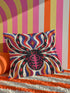 Square psychedelic folk art throw pillow cover printed to look like textured embroidery on a cream background. A large black spider with coral red and magenta markings on its legs and abdomen sits at the center, surrounded by rippling waves in blue violet, coral red, magenta, and golden yellow. Smooth flat 2D print, not raised stitching. Retro gothic Halloween maximalist decor for a sofa, armchair, or goth bedroom, made for spooky season.