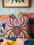 Square psychedelic folk art throw pillow cover printed to look like textured embroidery on a cream background. A large black spider with coral red and magenta markings on its legs and abdomen sits at the center, surrounded by rippling waves in blue violet, coral red, magenta, and golden yellow. Smooth flat 2D print, not raised stitching. Retro gothic Halloween maximalist decor for a sofa, armchair, or goth bedroom, made for spooky season.