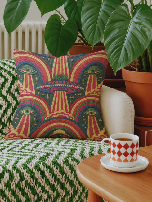 Square 70s psychedelic throw pillow cover printed to look like textured embroidery on a dark, starry navy background. Retro flying saucers and stylized alien eyes repeat across the design above rainbow arches, with small yellow stars and beams of light. Smooth flat 2D print, not raised stitching. Trippy, groovy, retro space age maximalist decor for a sofa, dorm bed, or reading nook, made for sci-fi fans and alien lovers.