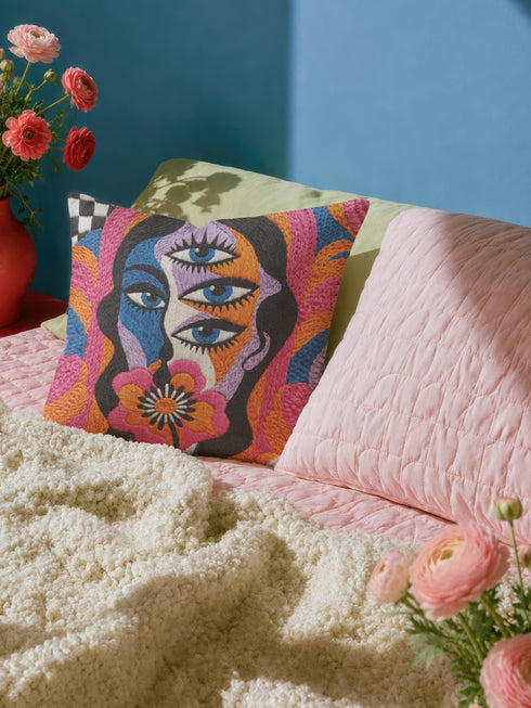 Square psychedelic throw pillow cover with a surreal weirdcore face printed to look like textured crewel embroidery. The face has a third eye and extra blue eyes, long dark hair, and a large pink and orange flower over the mouth, framed by radiating wavy stripes in hot pink, orange, and cobalt blue with black and white checkerboard corners. Smooth flat 2D print, not raised stitching. Maximalist 70s trippy decor for a sofa, armchair, or dorm bed.