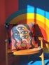 Square psychedelic throw pillow cover with a surreal weirdcore face printed to look like textured crewel embroidery. The face has a third eye and extra blue eyes, long dark hair, and a large pink and orange flower over the mouth, framed by radiating wavy stripes in hot pink, orange, and cobalt blue with black and white checkerboard corners. Smooth flat 2D print, not raised stitching. Maximalist 70s trippy decor for a sofa, armchair, or dorm bed.