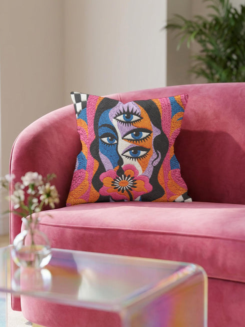 Square psychedelic throw pillow cover with a surreal weirdcore face printed to look like textured crewel embroidery. The face has a third eye and extra blue eyes, long dark hair, and a large pink and orange flower over the mouth, framed by radiating wavy stripes in hot pink, orange, and cobalt blue with black and white checkerboard corners. Smooth flat 2D print, not raised stitching. Maximalist 70s trippy decor for a sofa, armchair, or dorm bed.