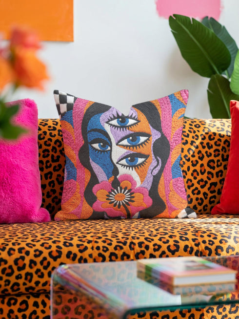Square psychedelic throw pillow cover with a surreal weirdcore face printed to look like textured crewel embroidery. The face has a third eye and extra blue eyes, long dark hair, and a large pink and orange flower over the mouth, framed by radiating wavy stripes in hot pink, orange, and cobalt blue with black and white checkerboard corners. Smooth flat 2D print, not raised stitching. Maximalist 70s trippy decor for a sofa, armchair, or dorm bed.