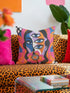 Square psychedelic throw pillow cover with a surreal weirdcore face printed to look like textured crewel embroidery. The face has a third eye and extra blue eyes, long dark hair, and a large pink and orange flower over the mouth, framed by radiating wavy stripes in hot pink, orange, and cobalt blue with black and white checkerboard corners. Smooth flat 2D print, not raised stitching. Maximalist 70s trippy decor for a sofa, armchair, or dorm bed.
