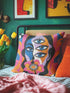Square psychedelic throw pillow cover with a surreal weirdcore face printed to look like textured crewel embroidery. The face has a third eye and extra blue eyes, long dark hair, and a large pink and orange flower over the mouth, framed by radiating wavy stripes in hot pink, orange, and cobalt blue with black and white checkerboard corners. Smooth flat 2D print, not raised stitching. Maximalist 70s trippy decor for a sofa, armchair, or dorm bed.