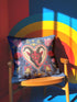 Square gothic Christmas throw pillow cover in a folk art design printed to look like textured crewel embroidery on a medium blue background. A red horned Krampus face with a long tongue sits inside a white and gold scalloped heart, surrounded by dark leafy vines, pink tulips, red carnations, gold bells tied with red bows, and small gold stars. Smooth flat 2D print, not raised stitching. Maximalist spooky holiday decor for a sofa, armchair, or goth bedroom.