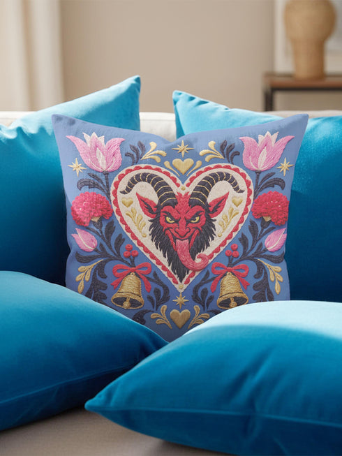 Square gothic Christmas throw pillow cover in a folk art design printed to look like textured crewel embroidery on a medium blue background. A red horned Krampus face with a long tongue sits inside a white and gold scalloped heart, surrounded by dark leafy vines, pink tulips, red carnations, gold bells tied with red bows, and small gold stars. Smooth flat 2D print, not raised stitching. Maximalist spooky holiday decor for a sofa, armchair, or goth bedroom.