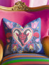 Square gothic Christmas throw pillow cover in a folk art design printed to look like textured crewel embroidery on a medium blue background. A red horned Krampus face with a long tongue sits inside a white and gold scalloped heart, surrounded by dark leafy vines, pink tulips, red carnations, gold bells tied with red bows, and small gold stars. Smooth flat 2D print, not raised stitching. Maximalist spooky holiday decor for a sofa, armchair, or goth bedroom.
