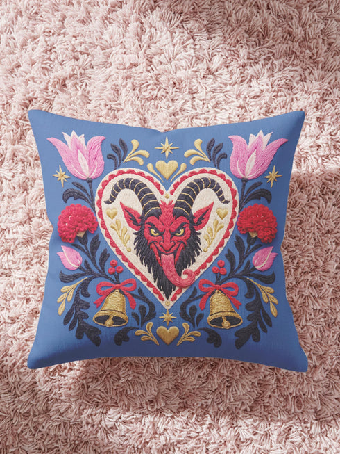 Square gothic Christmas throw pillow cover in a folk art design printed to look like textured crewel embroidery on a medium blue background. A red horned Krampus face with a long tongue sits inside a white and gold scalloped heart, surrounded by dark leafy vines, pink tulips, red carnations, gold bells tied with red bows, and small gold stars. Smooth flat 2D print, not raised stitching. Maximalist spooky holiday decor for a sofa, armchair, or goth bedroom.