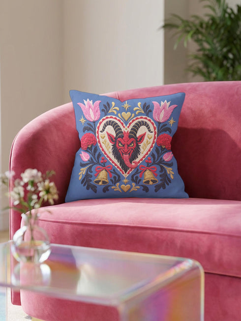 Square gothic Christmas throw pillow cover in a folk art design printed to look like textured crewel embroidery on a medium blue background. A red horned Krampus face with a long tongue sits inside a white and gold scalloped heart, surrounded by dark leafy vines, pink tulips, red carnations, gold bells tied with red bows, and small gold stars. Smooth flat 2D print, not raised stitching. Maximalist spooky holiday decor for a sofa, armchair, or goth bedroom.