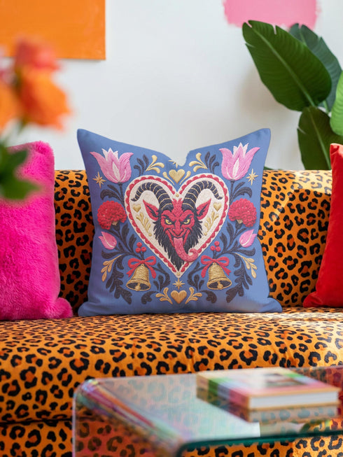 Square gothic Christmas throw pillow cover in a folk art design printed to look like textured crewel embroidery on a medium blue background. A red horned Krampus face with a long tongue sits inside a white and gold scalloped heart, surrounded by dark leafy vines, pink tulips, red carnations, gold bells tied with red bows, and small gold stars. Smooth flat 2D print, not raised stitching. Maximalist spooky holiday decor for a sofa, armchair, or goth bedroom.