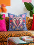 Square gothic Christmas throw pillow cover in a folk art design printed to look like textured crewel embroidery on a medium blue background. A red horned Krampus face with a long tongue sits inside a white and gold scalloped heart, surrounded by dark leafy vines, pink tulips, red carnations, gold bells tied with red bows, and small gold stars. Smooth flat 2D print, not raised stitching. Maximalist spooky holiday decor for a sofa, armchair, or goth bedroom.