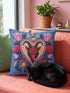 Square gothic Christmas throw pillow cover in a folk art design printed to look like textured crewel embroidery on a medium blue background. A red horned Krampus face with a long tongue sits inside a white and gold scalloped heart, surrounded by dark leafy vines, pink tulips, red carnations, gold bells tied with red bows, and small gold stars. Smooth flat 2D print, not raised stitching. Maximalist spooky holiday decor for a sofa, armchair, or goth bedroom.