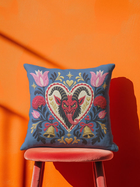 Square gothic Christmas throw pillow cover in a folk art design printed to look like textured crewel embroidery on a medium blue background. A red horned Krampus face with a long tongue sits inside a white and gold scalloped heart, surrounded by dark leafy vines, pink tulips, red carnations, gold bells tied with red bows, and small gold stars. Smooth flat 2D print, not raised stitching. Maximalist spooky holiday decor for a sofa, armchair, or goth bedroom.