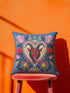 Square gothic Christmas throw pillow cover in a folk art design printed to look like textured crewel embroidery on a medium blue background. A red horned Krampus face with a long tongue sits inside a white and gold scalloped heart, surrounded by dark leafy vines, pink tulips, red carnations, gold bells tied with red bows, and small gold stars. Smooth flat 2D print, not raised stitching. Maximalist spooky holiday decor for a sofa, armchair, or goth bedroom.