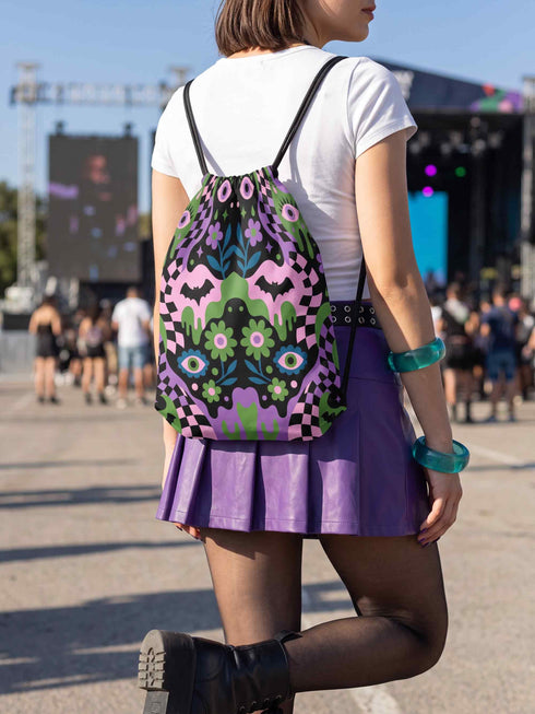 Drawstring backpack measuring 15 by 17 inches, made of 100% polyester with narrow shoulder straps and a drawstring closure. A symmetrical pastel goth print of bats, stylized eyes, flowers, and stars in a soft pastel palette sits on a black base, framed by purple and green checkerboard bands. Creepy cute, whimsigoth, kawaii goth style bag made for the gym, concerts, festivals, travel, school, and everyday carry.