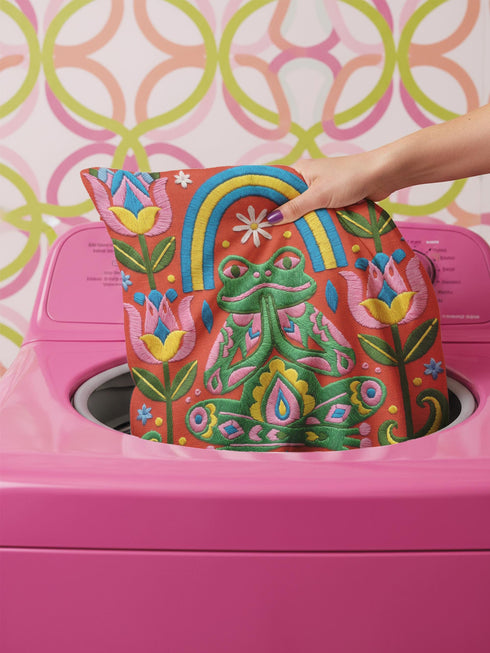 Square folk art throw pillow cover printed to look like textured crewel embroidery on a coral orange background. A green frog sits cross-legged in a meditation pose beneath a blue and yellow arch, surrounded by symmetrical pink flowers. Smooth flat 2D print, not raised stitching. Colorful, whimsical, maximalist dopamine decor for a yoga room, meditation corner, sofa, or reading nook, made for yoga lovers and frog fans.