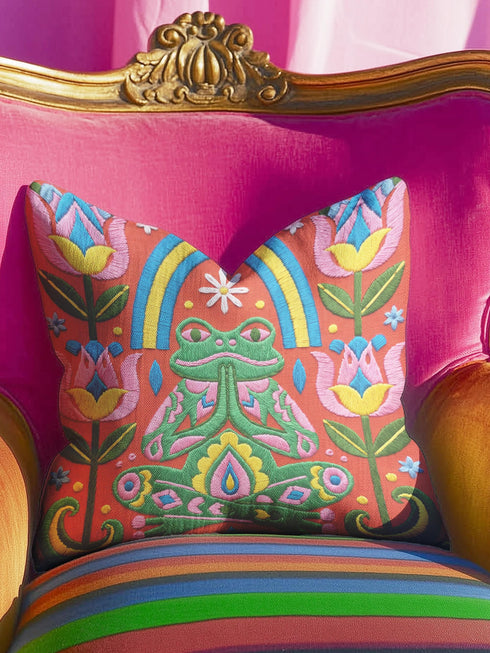 Square folk art throw pillow cover printed to look like textured crewel embroidery on a coral orange background. A green frog sits cross-legged in a meditation pose beneath a blue and yellow arch, surrounded by symmetrical pink flowers. Smooth flat 2D print, not raised stitching. Colorful, whimsical, maximalist dopamine decor for a yoga room, meditation corner, sofa, or reading nook, made for yoga lovers and frog fans.
