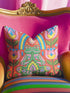 Square folk art throw pillow cover printed to look like textured crewel embroidery on a coral orange background. A green frog sits cross-legged in a meditation pose beneath a blue and yellow arch, surrounded by symmetrical pink flowers. Smooth flat 2D print, not raised stitching. Colorful, whimsical, maximalist dopamine decor for a yoga room, meditation corner, sofa, or reading nook, made for yoga lovers and frog fans.
