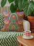 Square folk art throw pillow cover printed to look like textured crewel embroidery on a coral orange background. A green frog sits cross-legged in a meditation pose beneath a blue and yellow arch, surrounded by symmetrical pink flowers. Smooth flat 2D print, not raised stitching. Colorful, whimsical, maximalist dopamine decor for a yoga room, meditation corner, sofa, or reading nook, made for yoga lovers and frog fans.
