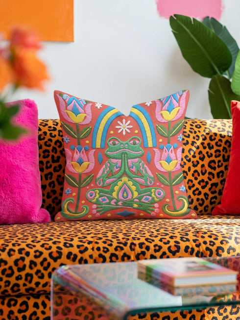 Square folk art throw pillow cover printed to look like textured crewel embroidery on a coral orange background. A green frog sits cross-legged in a meditation pose beneath a blue and yellow arch, surrounded by symmetrical pink flowers. Smooth flat 2D print, not raised stitching. Colorful, whimsical, maximalist dopamine decor for a yoga room, meditation corner, sofa, or reading nook, made for yoga lovers and frog fans.