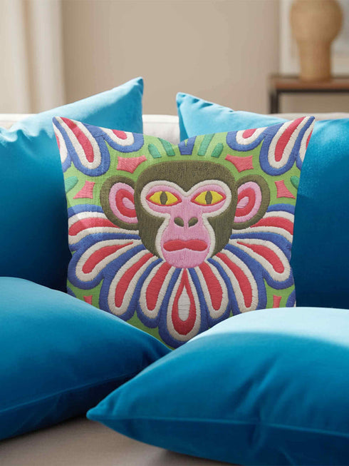 Square folk art throw pillow cover printed to look like thick crewel yarn embroidery. A bold, whimsical monkey face in olive green and pink stares out with intense yellow eyes, framed by a retro scalloped floral collar. Smooth flat 2D print, not raised stitching. Clowncore, junglecore, and weirdcore maximalist decor with eclectic, quirky charm for a sofa, accent chair, or bedroom, made for animal lovers and alt renters.