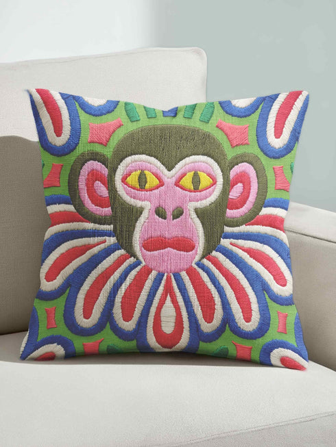 Square folk art throw pillow cover printed to look like thick crewel yarn embroidery. A bold, whimsical monkey face in olive green and pink stares out with intense yellow eyes, framed by a retro scalloped floral collar. Smooth flat 2D print, not raised stitching. Clowncore, junglecore, and weirdcore maximalist decor with eclectic, quirky charm for a sofa, accent chair, or bedroom, made for animal lovers and alt renters.