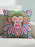 Square folk art throw pillow cover printed to look like thick crewel yarn embroidery. A bold, whimsical monkey face in olive green and pink stares out with intense yellow eyes, framed by a retro scalloped floral collar. Smooth flat 2D print, not raised stitching. Clowncore, junglecore, and weirdcore maximalist decor with eclectic, quirky charm for a sofa, accent chair, or bedroom, made for animal lovers and alt renters.