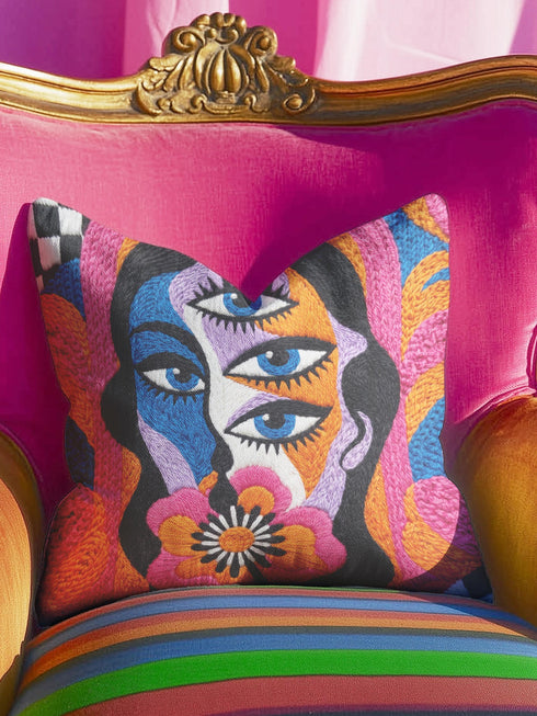 Square psychedelic throw pillow cover with a surreal weirdcore face printed to look like textured crewel embroidery. The face has a third eye and extra blue eyes, long dark hair, and a large pink and orange flower over the mouth, framed by radiating wavy stripes in hot pink, orange, and cobalt blue with black and white checkerboard corners. Smooth flat 2D print, not raised stitching. Maximalist 70s trippy decor for a sofa, armchair, or dorm bed.
