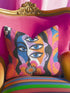 Square psychedelic throw pillow cover with a surreal weirdcore face printed to look like textured crewel embroidery. The face has a third eye and extra blue eyes, long dark hair, and a large pink and orange flower over the mouth, framed by radiating wavy stripes in hot pink, orange, and cobalt blue with black and white checkerboard corners. Smooth flat 2D print, not raised stitching. Maximalist 70s trippy decor for a sofa, armchair, or dorm bed.