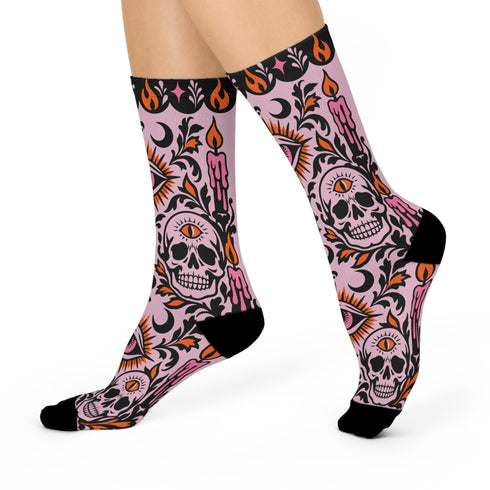 Pair of crew length socks with an all-over pastel goth print on a pastel pink background. Black-outlined human skulls with small orange eyes on their foreheads, all-seeing eyes with orange rays, crescent moons, curling leaves, and pink diamond stars sit in a symmetrical dark damask layout. Black cuff with a scalloped edge, orange flames, and pink flourishes. Reinforced black heel and toe, striped ribbed cuff. Western goth tattoo flash style.