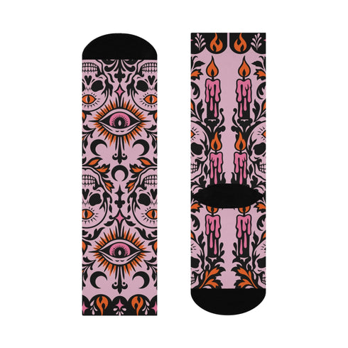Pair of crew length socks with an all-over pastel goth print on a pastel pink background. Black-outlined human skulls with small orange eyes on their foreheads, all-seeing eyes with orange rays, crescent moons, curling leaves, and pink diamond stars sit in a symmetrical dark damask layout. Black cuff with a scalloped edge, orange flames, and pink flourishes. Reinforced black heel and toe, striped ribbed cuff. Western goth tattoo flash style.