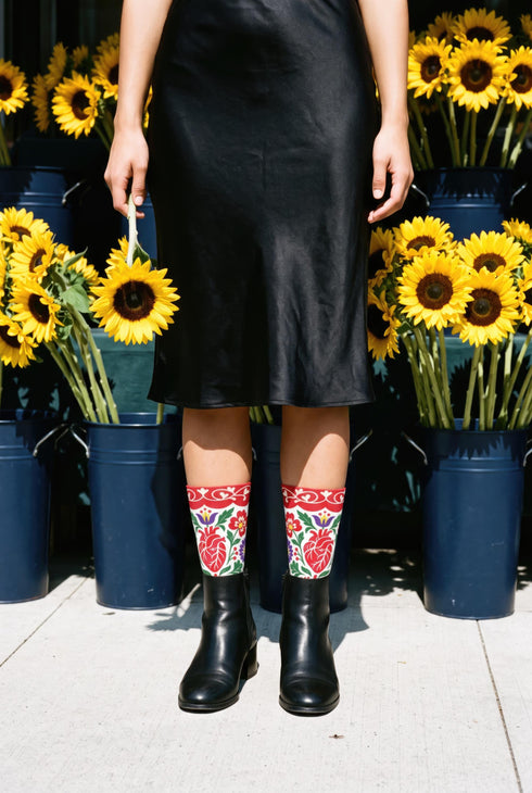 Pair of crew length socks with an all-over folk art print on a cream background. Red anatomical hearts stack vertically between symmetrical floral sprays with fine cream vessel lines, curling green stems and leaves, red flowers, purple tulip-shaped blooms, and small yellow accents. Red cuffs carry cream scrolling vines and small hearts above a scalloped border. Reinforced black heel and toe, striped ribbed cuff. Maximalist medical gift socks.